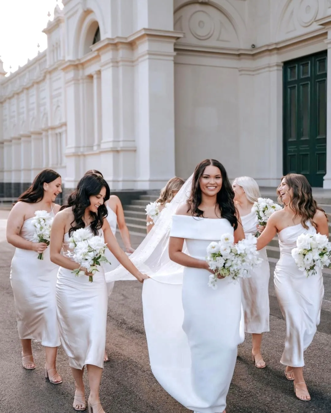 Bride and bridesmaids in white walking before a grand columned building, all bronzed