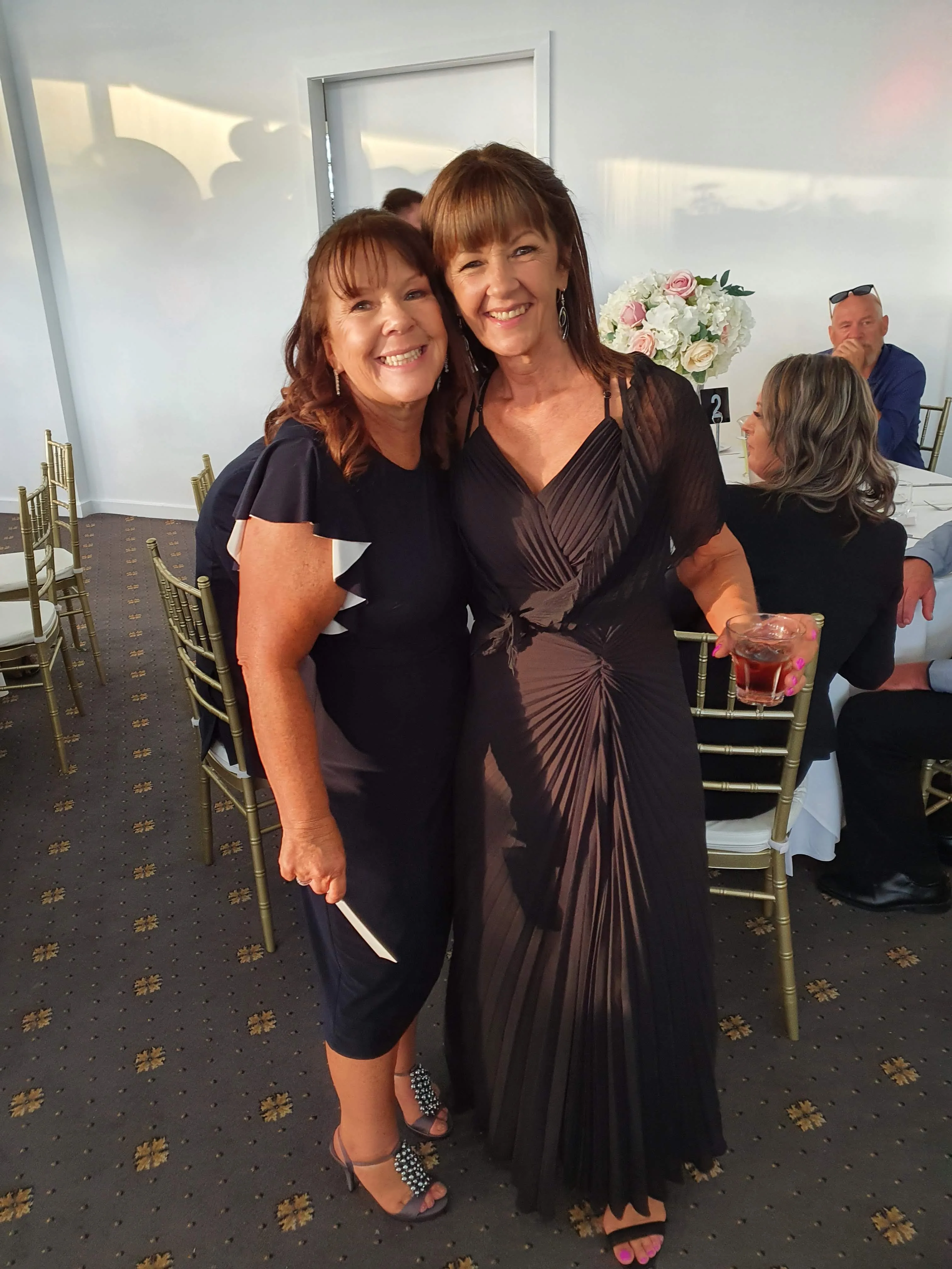 Two clients in evening dress inside a grand reception venue with gold chairs, both glowing
