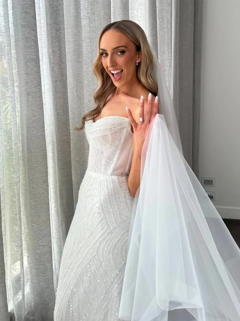 Bride in a beaded gown by sheer curtains, bronzed shoulders and arms glowing