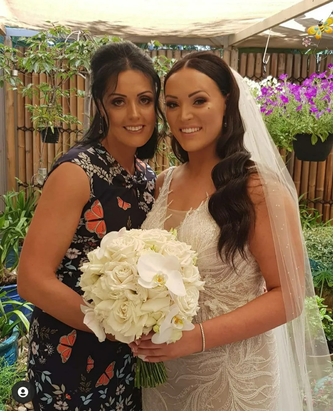 Bride and a guest in a leafy garden, both bronzed and radiant